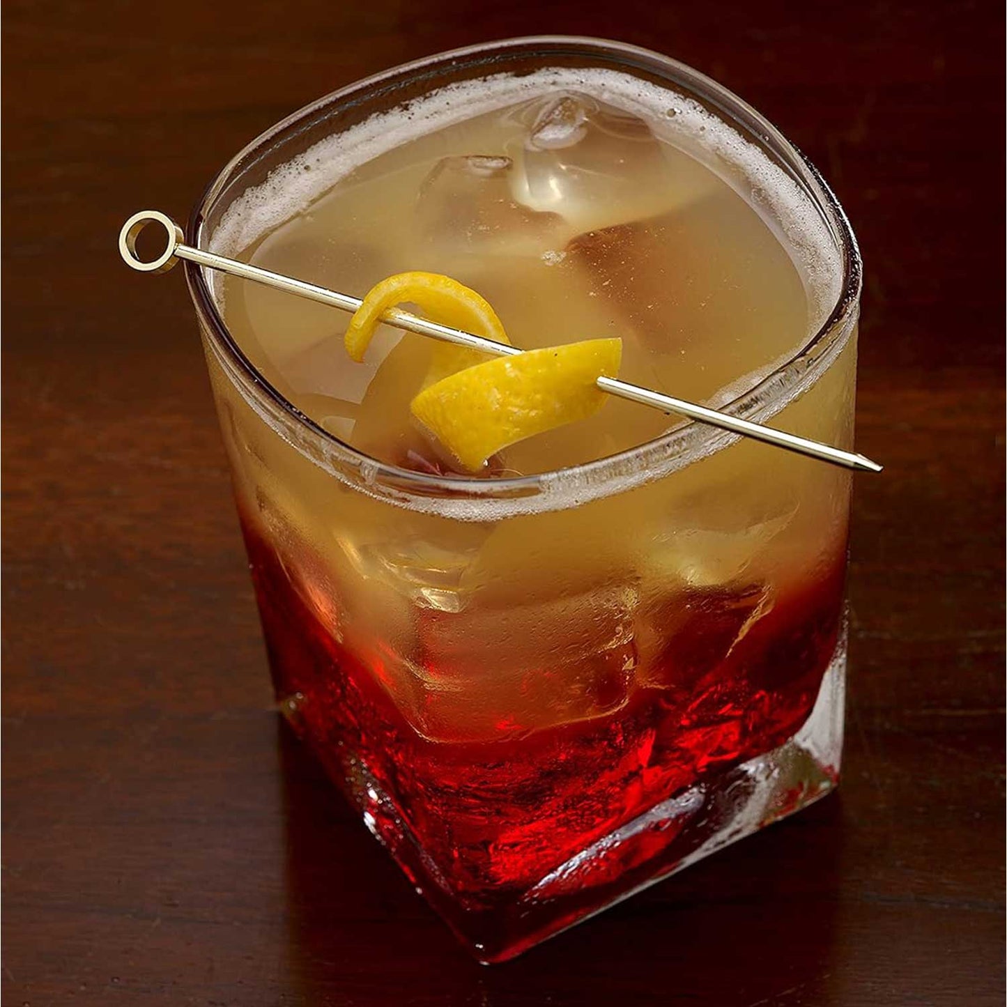 An amber and red layered cocktail, served over ice in a square rocks glass. A yellow citrus peel is speared across the top of the glass by a 4.5 inch Gold Plated Barfly Cocktail Pick, with a Circle top