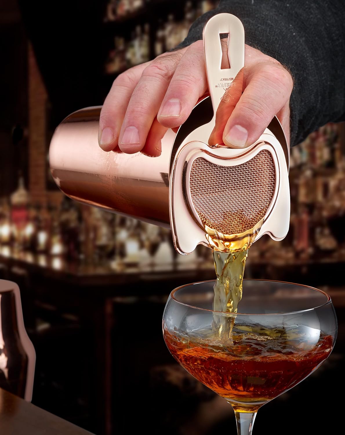 A bartender pouring a cocktail from a shaker with a Copper plated finished Barfly Fine Mesh Spring Strainer on a glass with ice