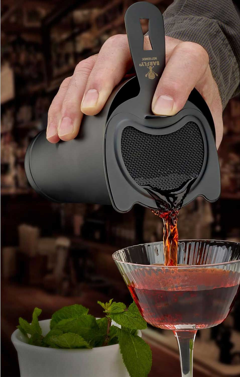 A bartender pours a red colored cocktail drink from a cocktail shaker with a Matte Black Barfly Fine Mesh Spring Strainer into a glass with a blurred bar background