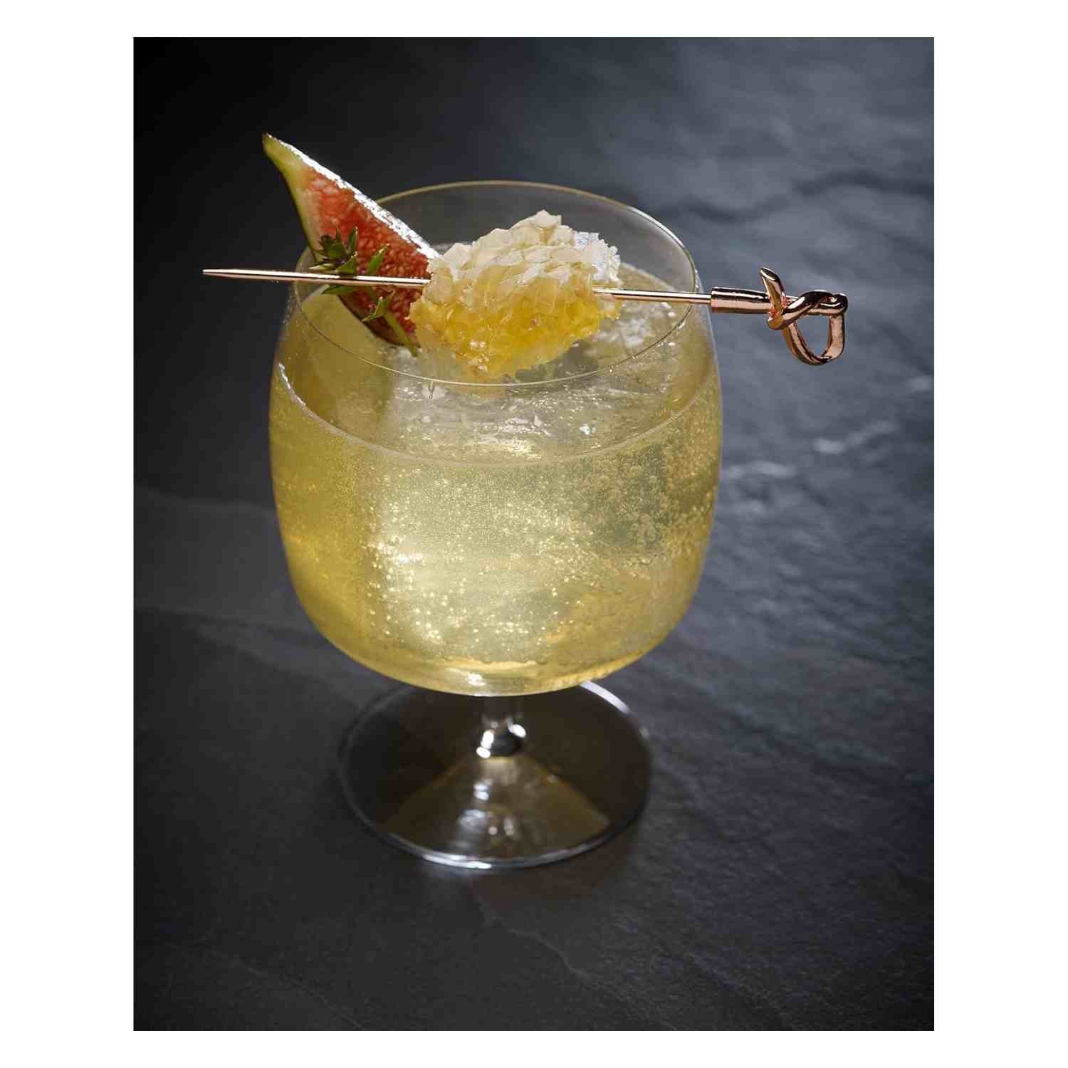 A sparkling, pale yellow cocktail in a wide-bowled stemmed glass, garnished with a slice of fig and a piece of honeycomb. The garnish is skewered on a copper-plated cocktail pick with a 4.5 inch Copper Plated Barfly Cocktail Pick, with a bamboo knot top.