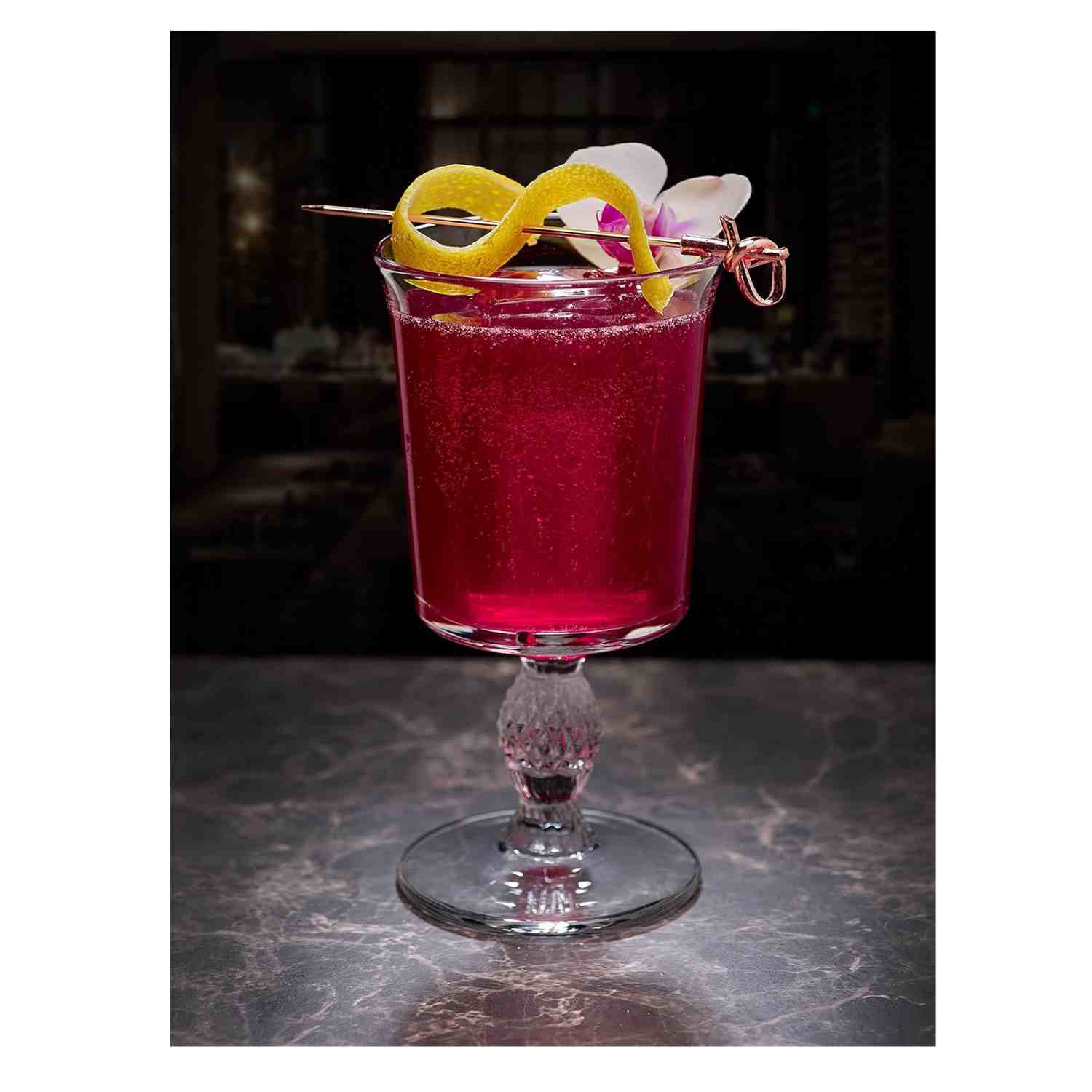 A deep magenta cocktail in a stemmed glass with a decorative, diamond-cut base. The drink is garnished with a lemon twist and a white orchid flower, skewered on a 4.5 inch Copper Plated Barfly Cocktail Pick, with a bamboo knot top.