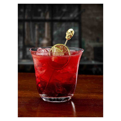 A bright, cranberry-red cocktail with ice and a slice of dried lime, served in a clear, fluted rocks glass on a dark wooden table. The garnish is held by a 4.5 inch Gold Plated Barfly Cocktail Pick, with a skull top