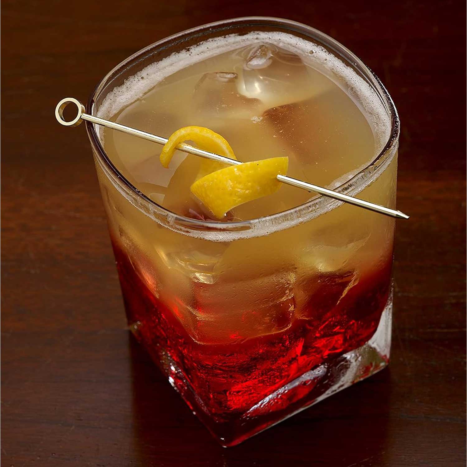 An amber and red layered cocktail, served over ice in a square rocks glass. A yellow citrus peel is speared across the top of the glass by a 4.5 inch Gold Plated Barfly Cocktail Pick, with a Circle top