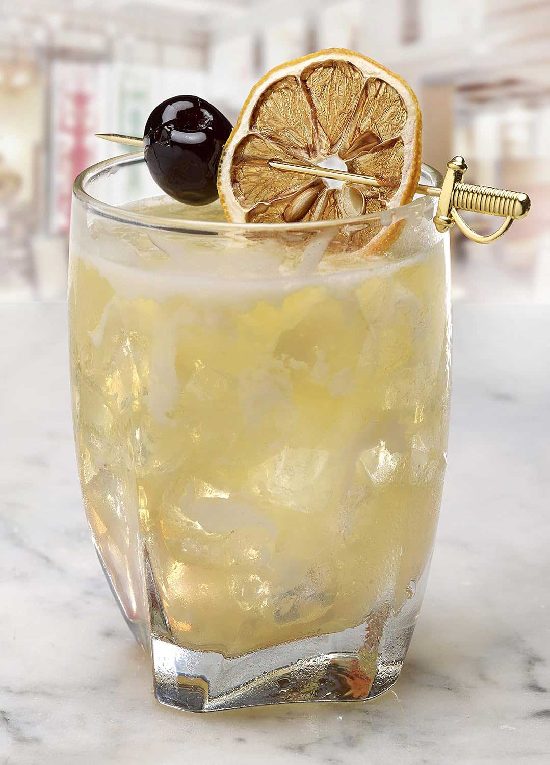 A yellow colored cocktail with ice in a glass on a marble surface. A lemon wheel and cherry garnish speared across the top of the glass by a 4.5 inch Gold Plated Barfly Cocktail Pick, with a Sword top. 