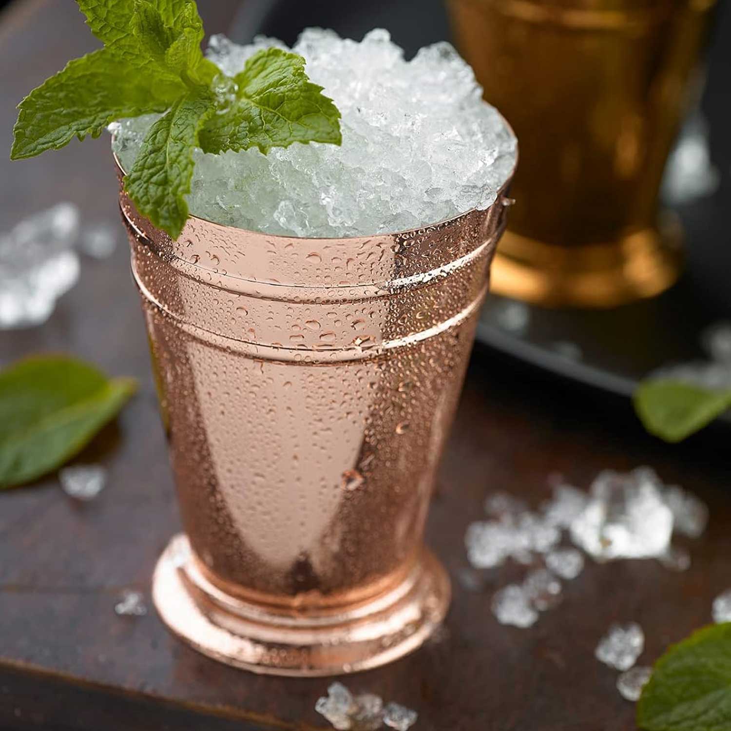 A Copper Plated Barfly Deluxe Julep Cup 13.5oz with crushed ice and mint leaves on a table with blurred background