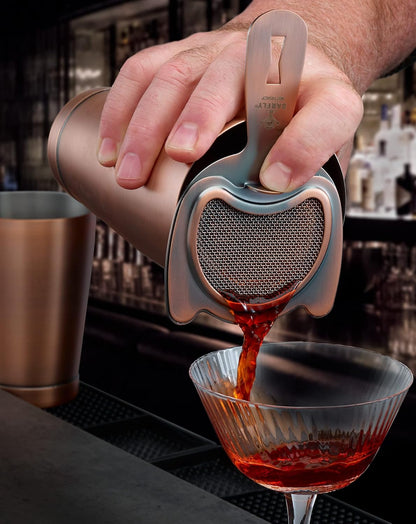 A bartender pouring a cocktail from a shaker with an Antique Copper finished Barfly Fine Mesh Spring Strainer on a glass