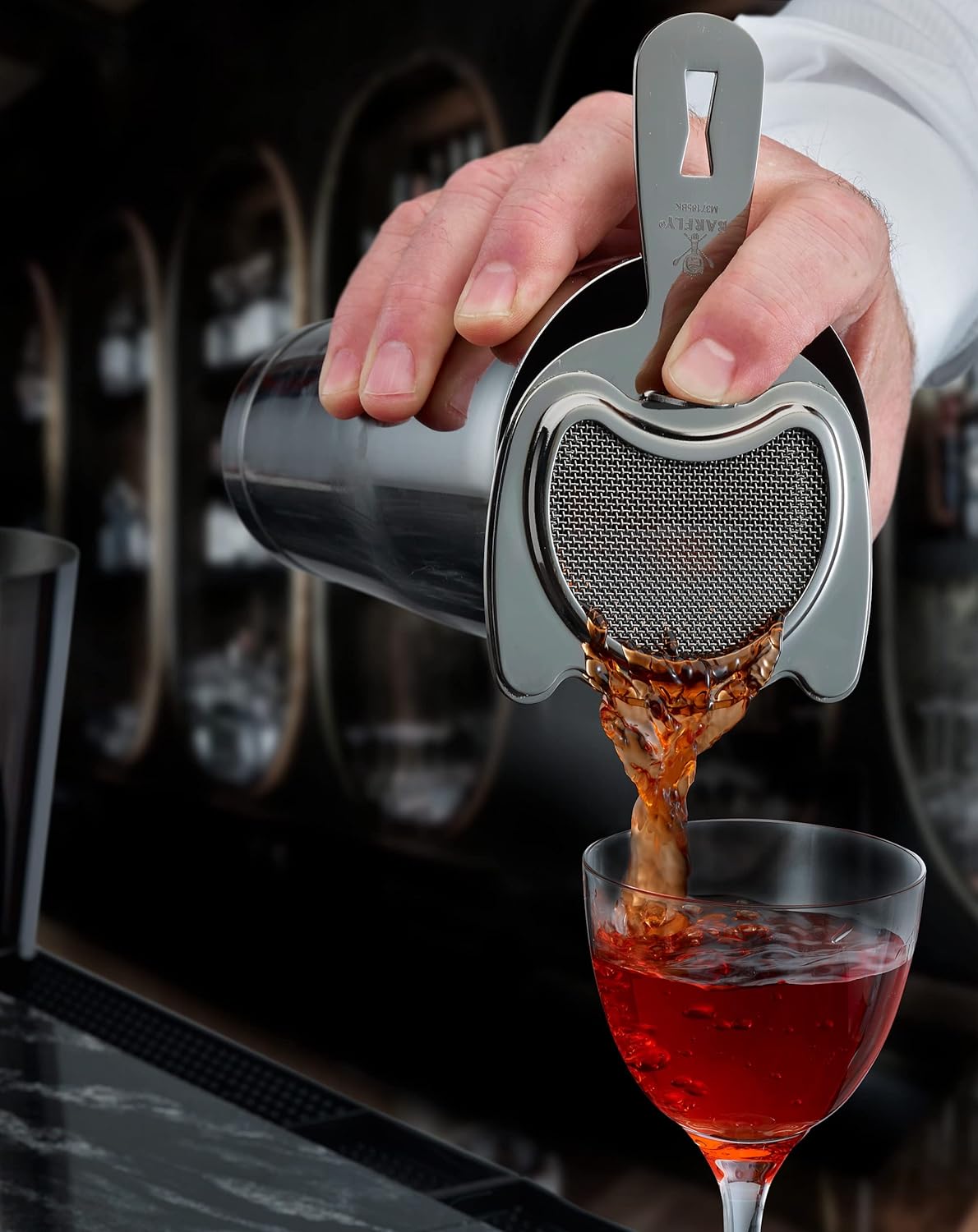 A bartender pours a red colored cocktail drink from a cocktail shaker with a Black Barfly Fine Mesh Spring Strainer into a glass with a blurred bar background