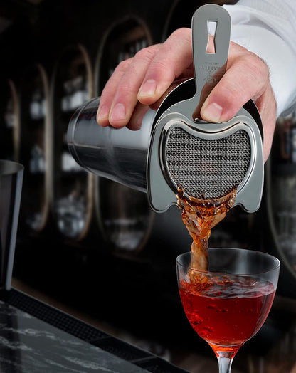 A bartender pours a red colored cocktail drink from a cocktail shaker with a Black Barfly Fine Mesh Spring Strainer into a glass with a blurred bar background