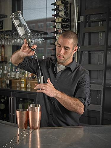 Man preparing a cocktail using the Stainless Barfly Japanese Style Jigger, 1 oz x 2 oz, in a bar setting with bottles and glasses in the background.