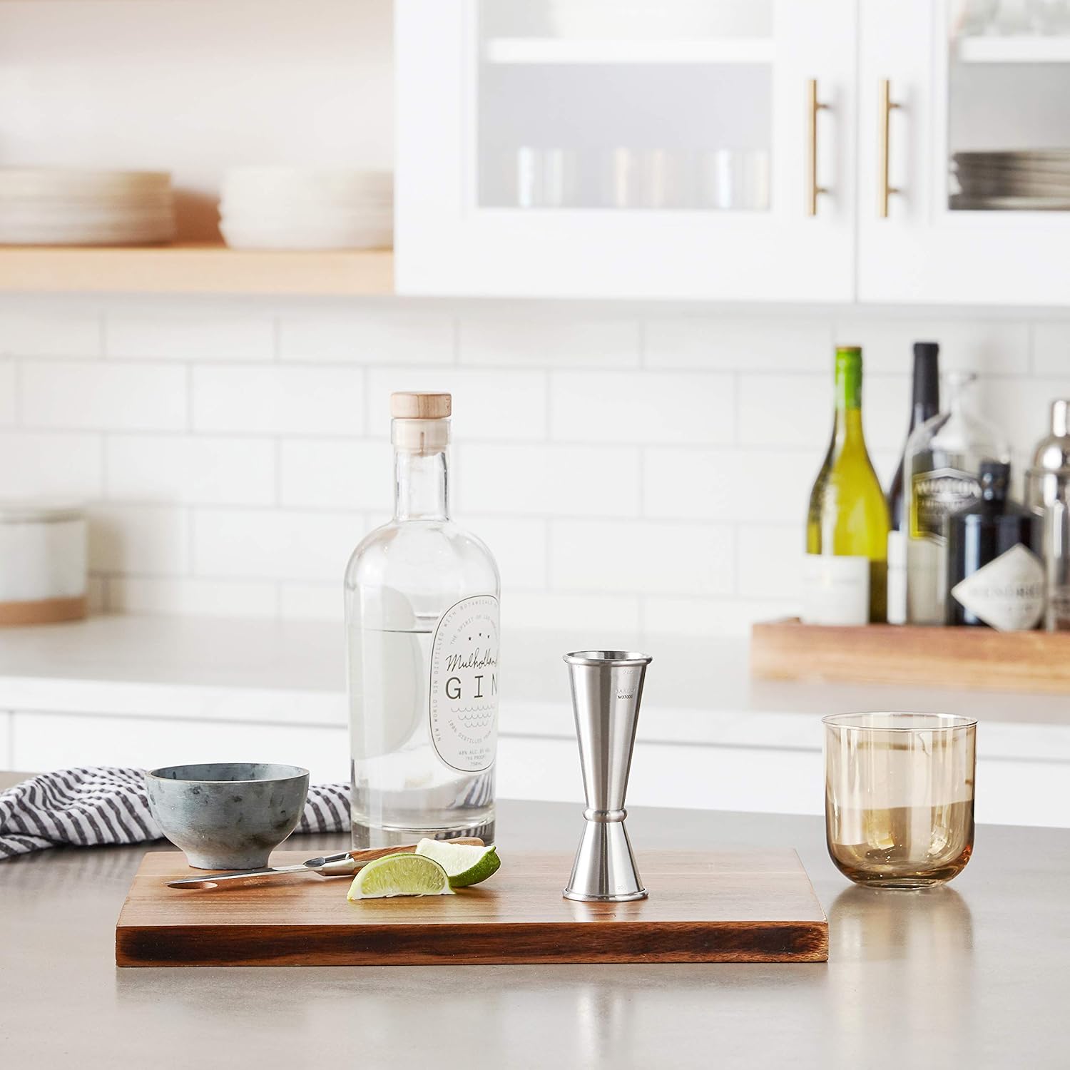 Bottle of gin, lime, A Stainless Barfly Japanese Style Jigger, 1 oz x 2 oz, and other bar tools on a wooden cutting board in a kitchen setting.