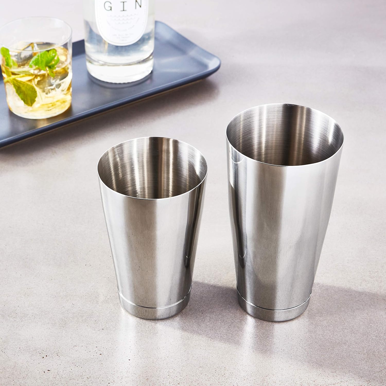 A barfly stainless steel shaker cocktail tin set of 18 oz and 28 oz on a light surface with a blurred background showing a cocktail and a bottle of gin.