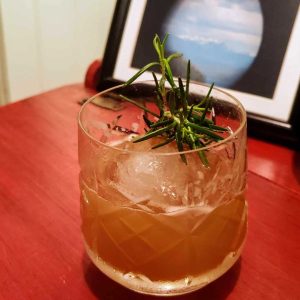 A cocktail in a rocks glass with ice cubes, garnished with a sprig of rosemary.