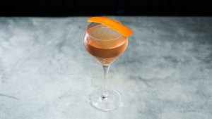 A cocktail in a coupe glass with ice, garnished with an orange peel.