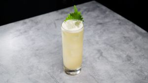 A tall glass filled with a light yellow cocktail, topped with ice cubes and garnished with a shiso leaf.